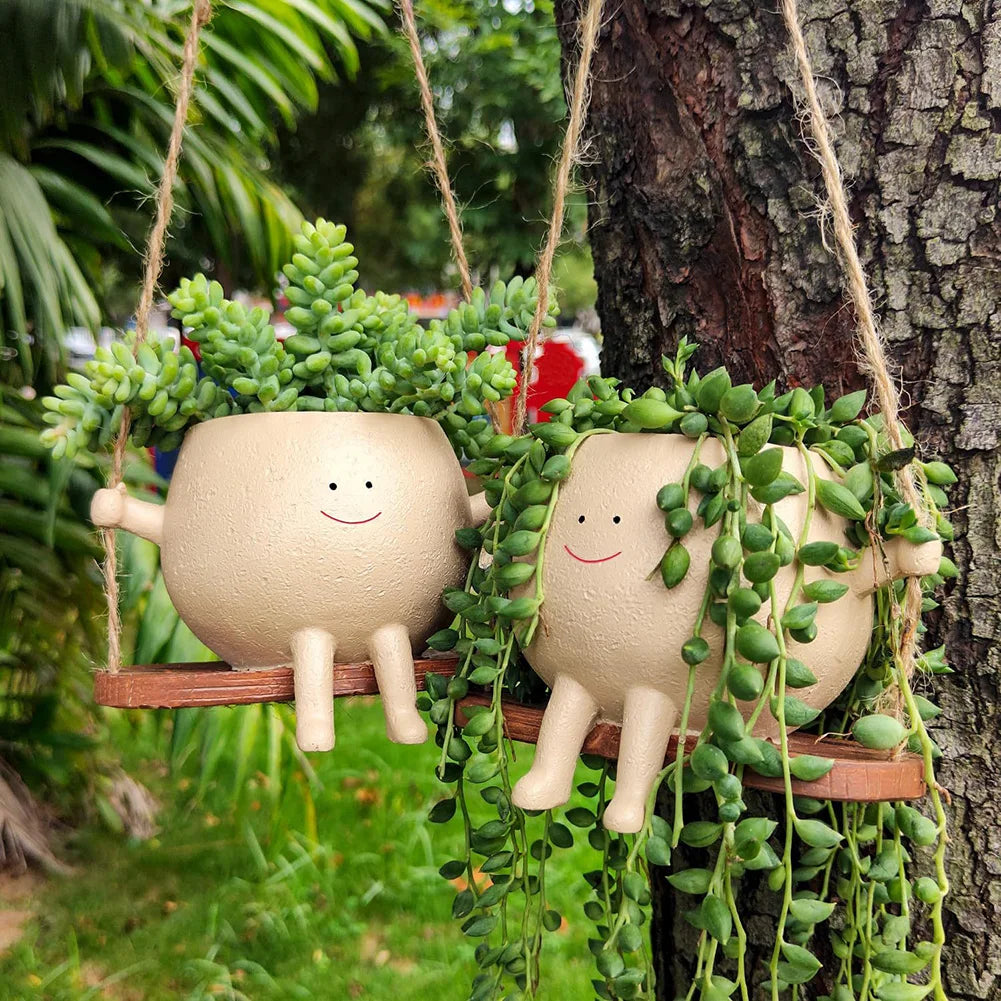 Two smiling face planters with succulents hanging in a garden, one trailing